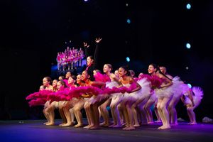 Dancers in pink costumes perform a synchronized dance routine on a dark stage with dramatic lighting effects.