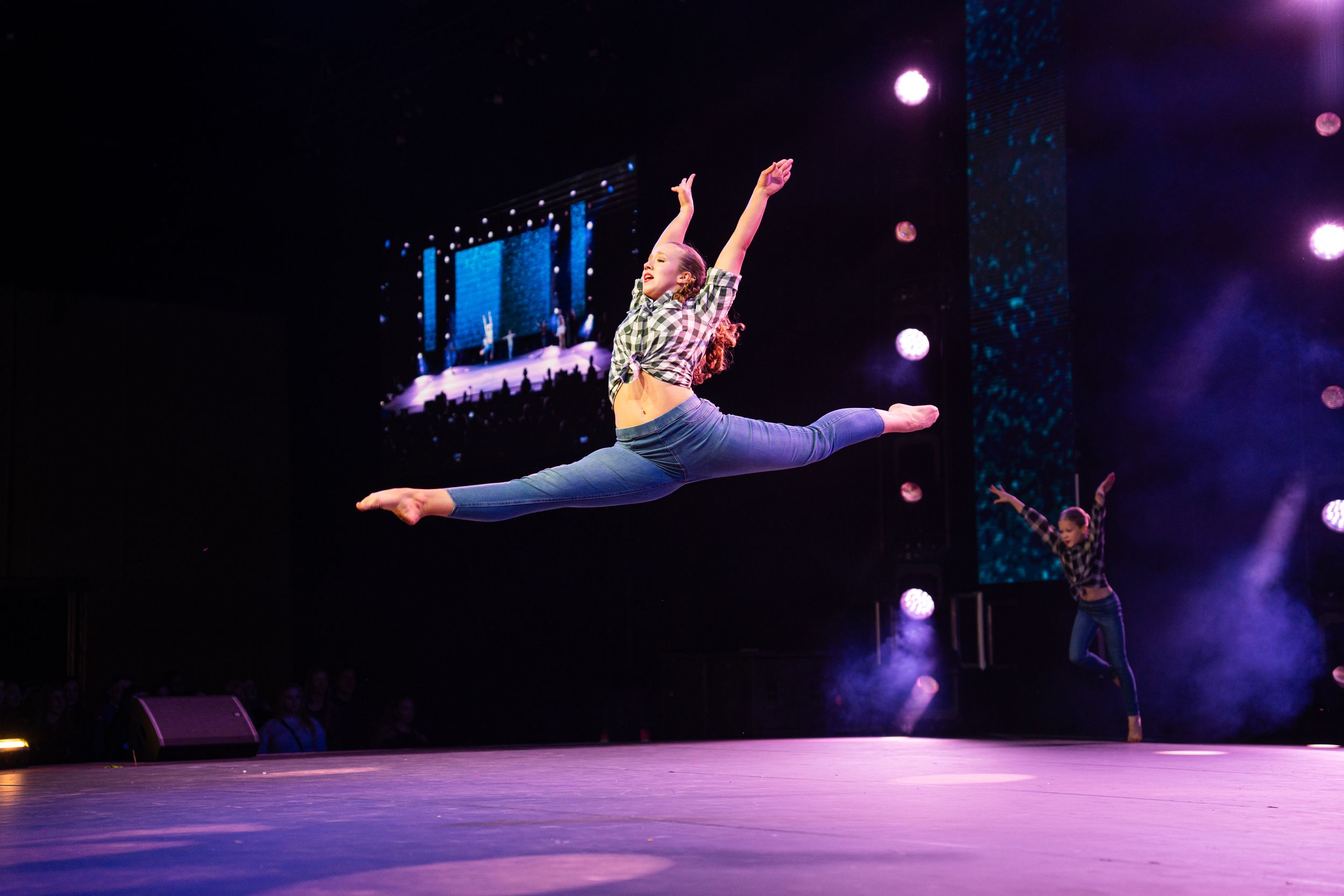A dancer performs a dramatic split leap in mid-air on stage with purple lighting and video screens in the background.