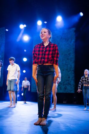 A performer in a red plaid shirt and dark pants stands on a blue-lit stage during a theatrical performance.