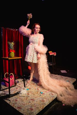 A performer in a floral dress and dramatic feather boa poses on stage with a pineapple prop. Theatre photography in Surrey.