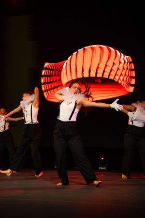 A sequence of dance performers in black pants moving with a glowing orange accordion-like prop on a dark stage.