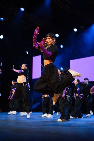 Dancers perform energetic hip-hop choreography on stage with dramatic blue lighting and matching black outfits.