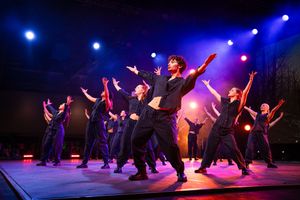 Dancers perform a synchronized routine on stage with dramatic blue and red lighting effects in a theatrical production.