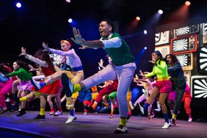 Energetic dancers perform synchronized jumps in colorful outfits on stage with vibrant lighting and theatrical backdrop.