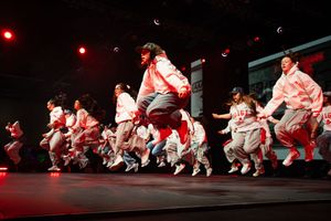 Dancers in white costumes perform a synchronized crouching movement on stage under dramatic red lighting.
