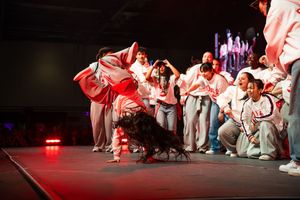 Performers in white costumes create dynamic movements during a stage performance with dramatic red lighting.