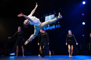 Dancers perform dynamic leaps and acrobatic moves on a dark stage illuminated by blue spotlights.