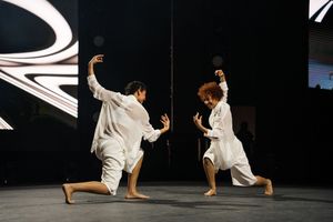 Martial artists in white uniforms demonstrate synchronized combat moves on a dark stage.