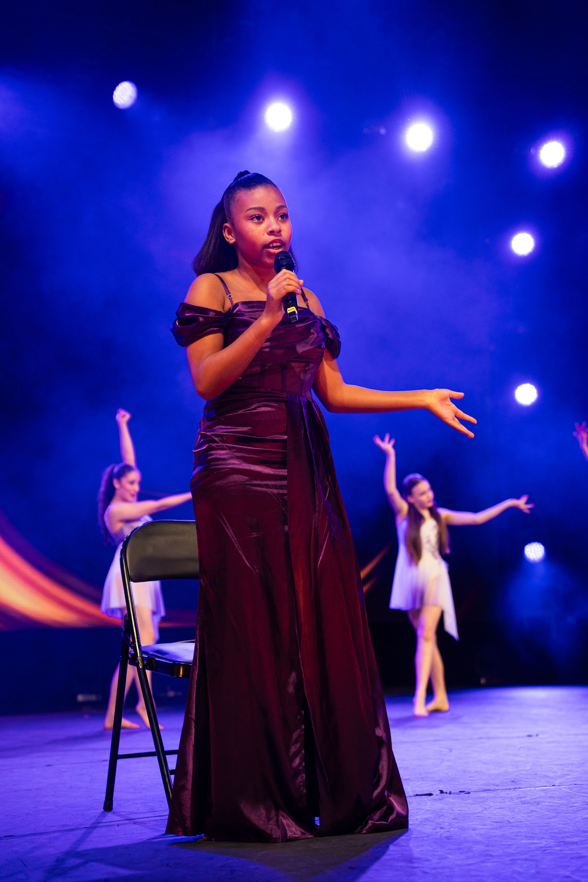 A performer in a burgundy velvet dress sits on a chair on stage with dramatic blue lighting and spotlights.