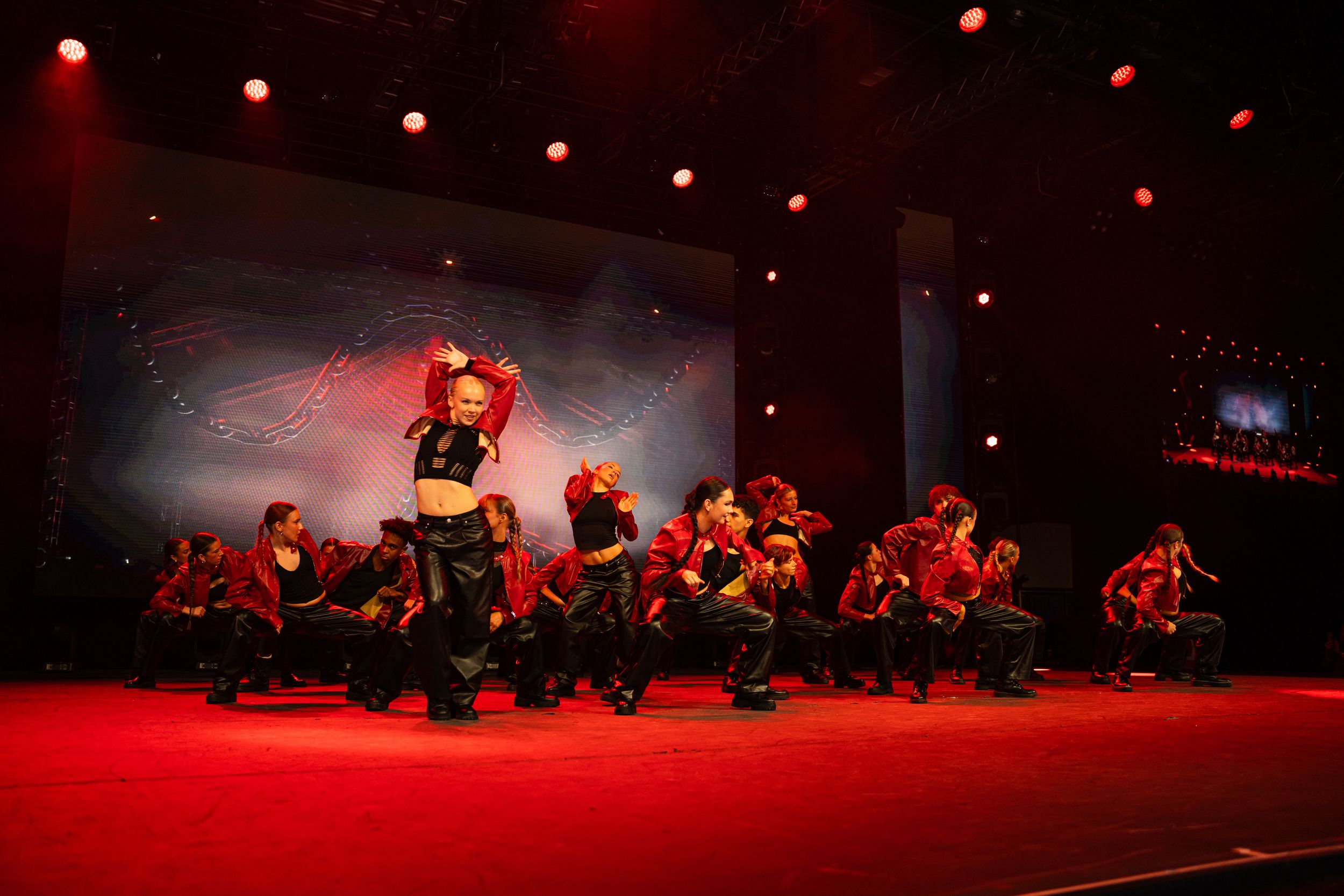 Dramatic dance performance on dark stage with red lighting and multiple performers in silhouette creating dynamic movements.