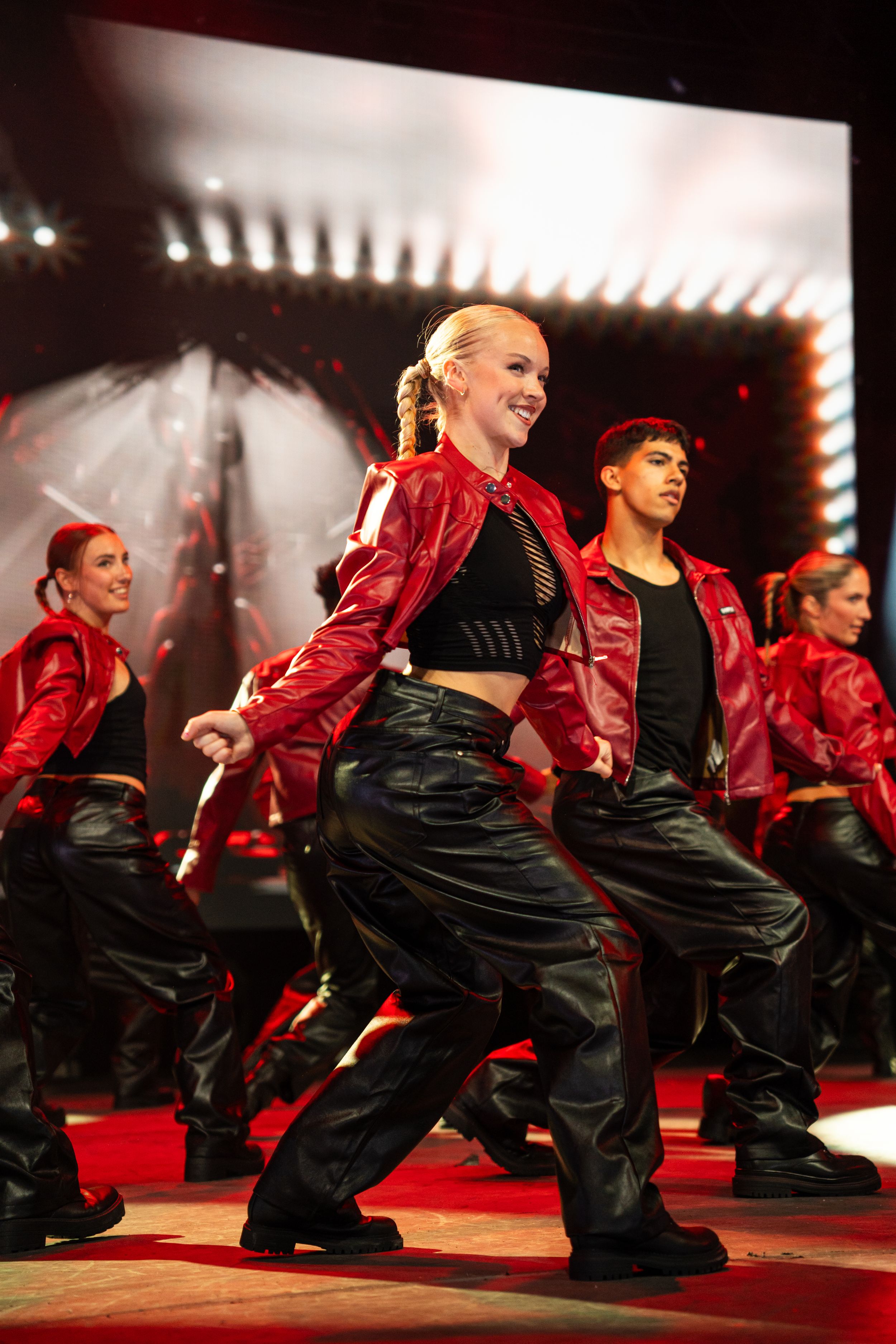 Dance performers in red jackets and black pants perform a synchronized crouching move during a stage performance.