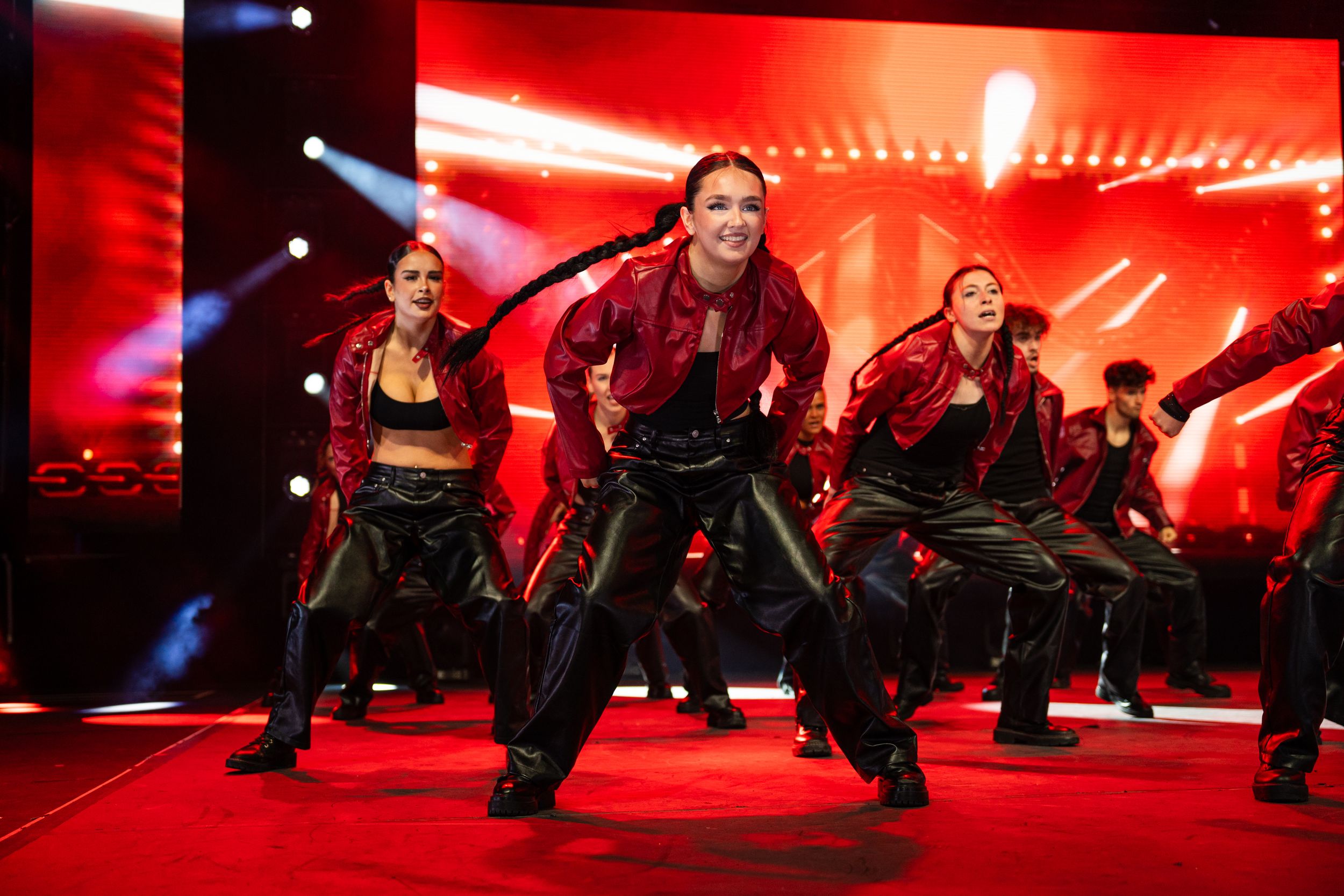 Dancers in black outfits perform a synchronized dance routine against a dramatic red-lit stage background.