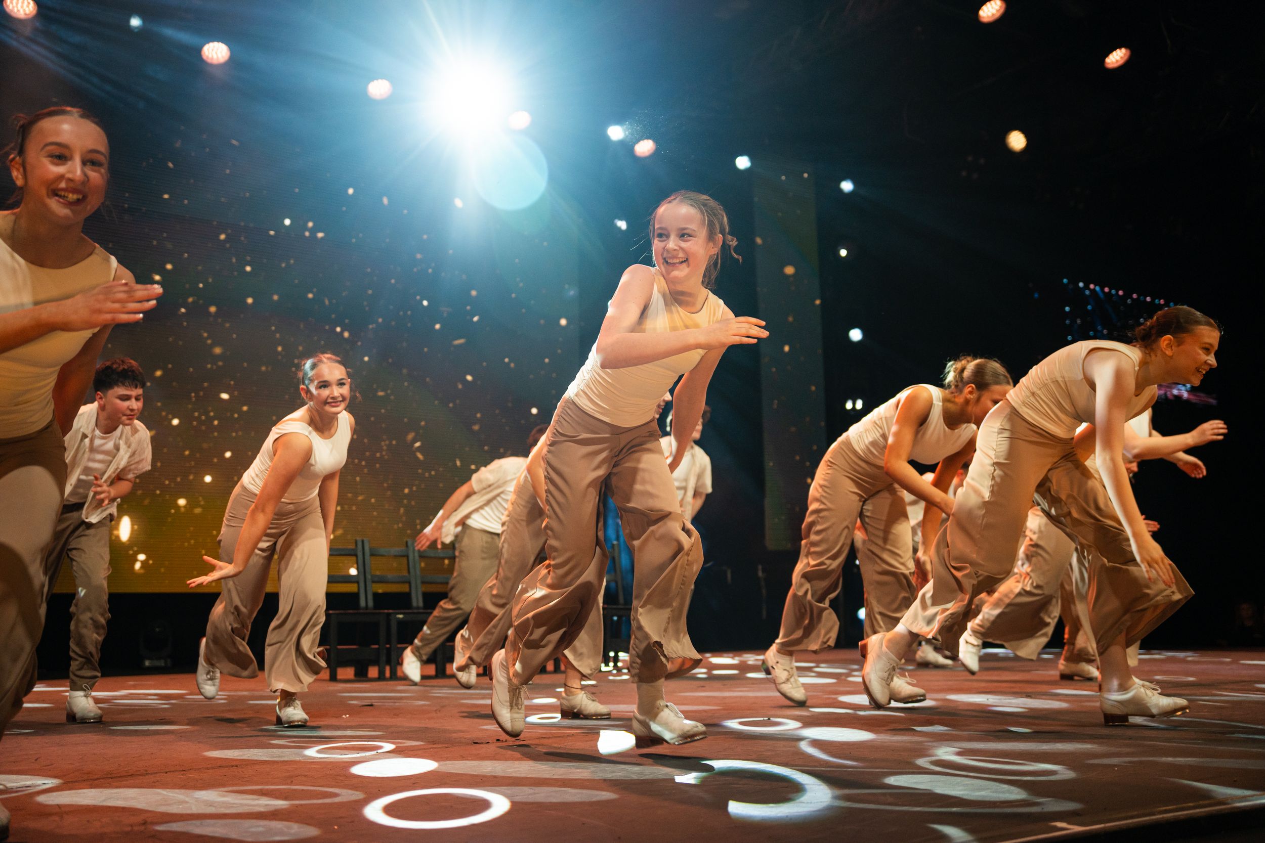 Dancers in white costumes perform an energetic choreographed sequence on a dark stage under bright spotlights.