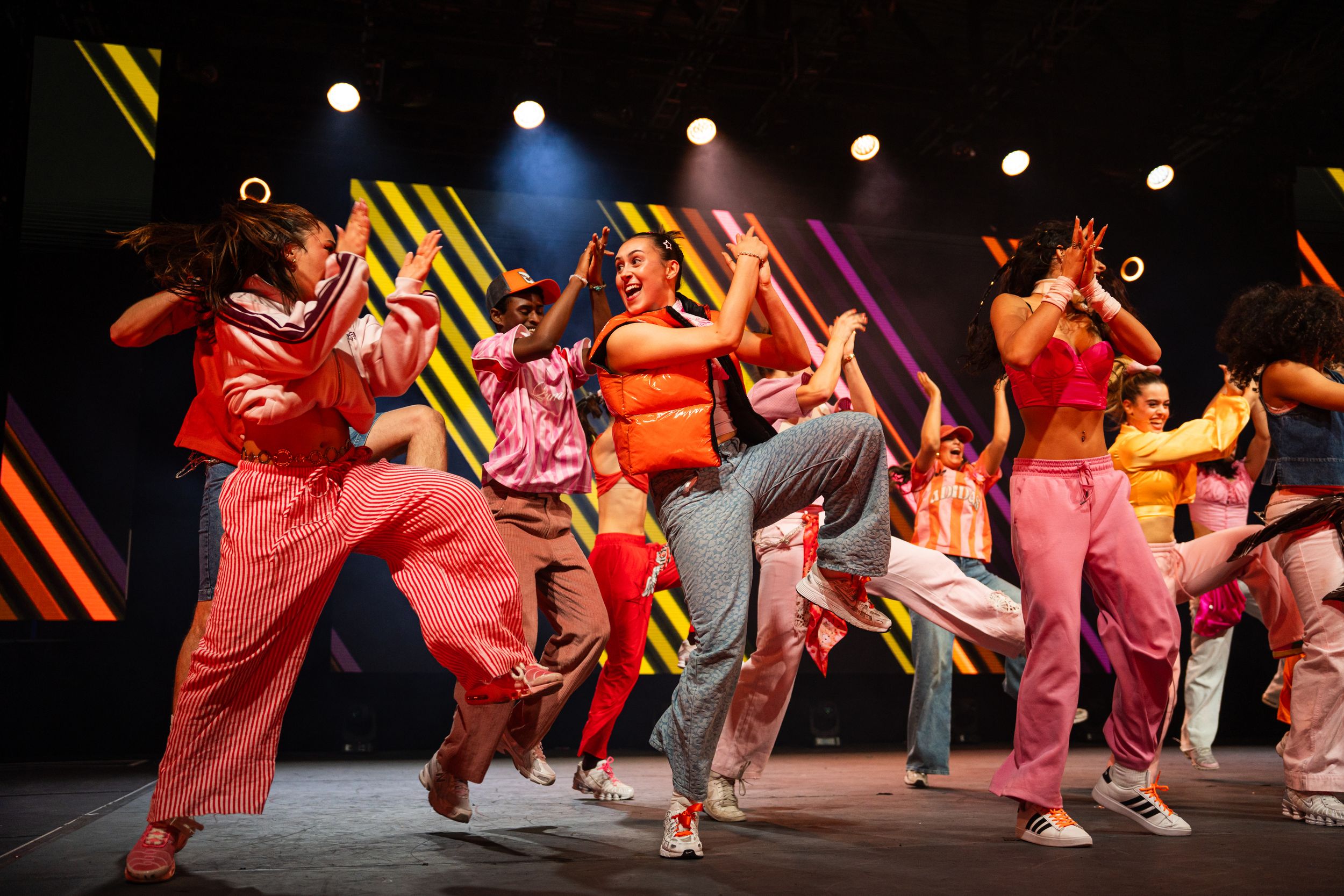 Dancers in vibrant orange and red costumes perform energetic choreography on a dark stage under spotlights.