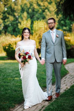 A bride in a lace gown and groom in a gray suit hold hands while walking along a garden path on their wedding day.