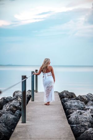A figure in a flowing white dress walks along a concrete pier with rocky breakwater and serene ocean views at sunset.
