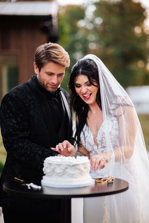 A bride and groom share a joyful moment while cutting their white wedding cake together at an outdoor reception.
