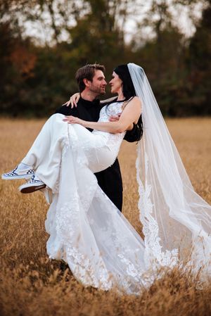 A romantic wedding photo in a golden field shows a couple embracing as the groom carries the bride in her lace-trimmed gown.