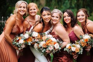 Wedding party poses with orange and white floral bouquets wearing burgundy and rust colored dresses.