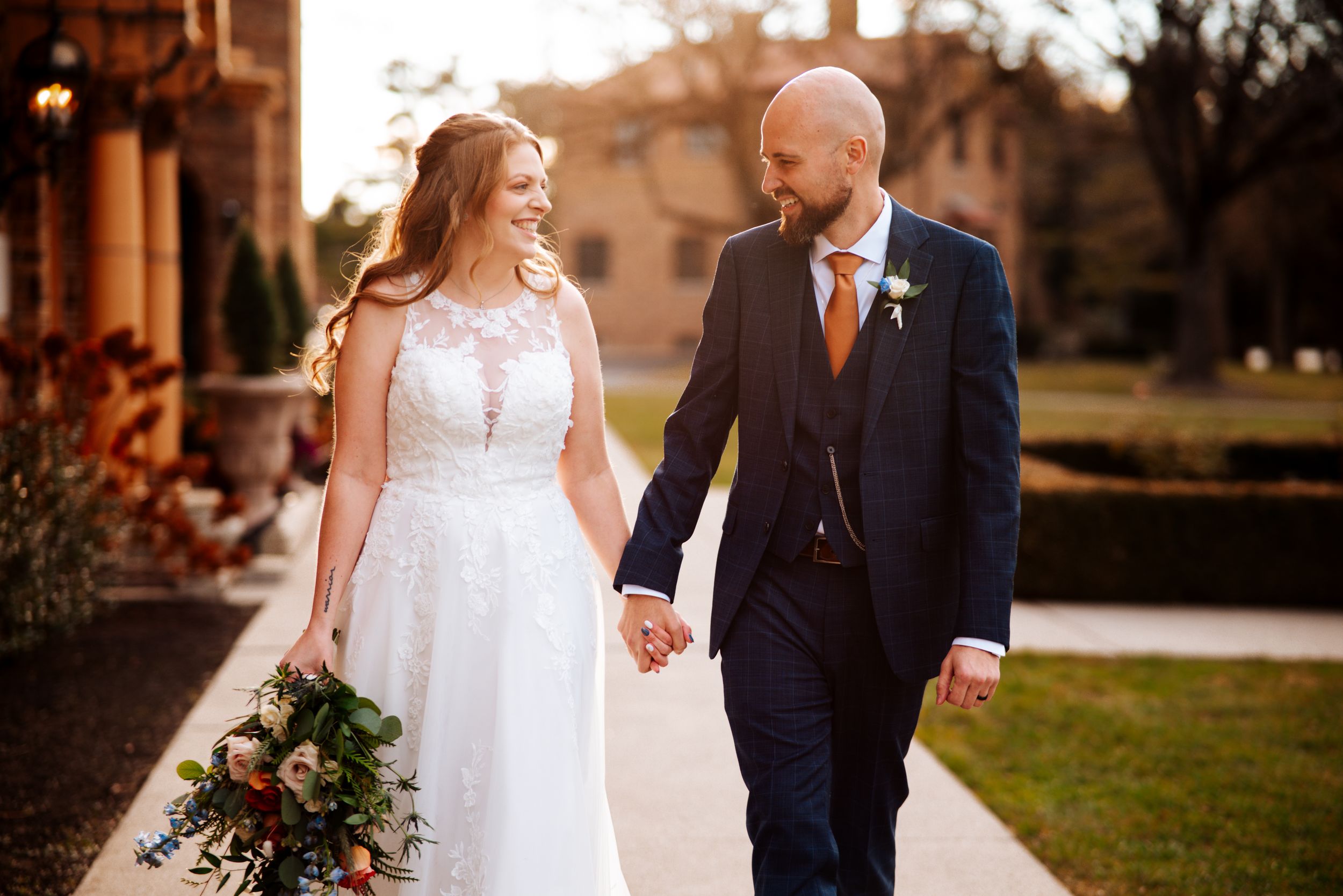 A romantic outdoor wedding photo shoot with a couple in formal attire holding hands at sunset.