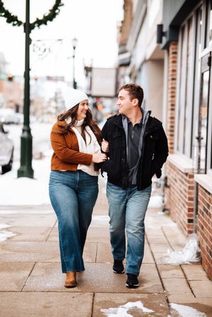 A couple walks together holding hands along a snowy downtown sidewalk with decorative lamp posts.