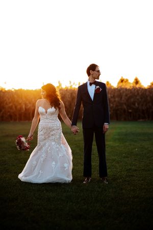 A couple in wedding attire holds hands while standing in a field at sunset, silhouetted against the golden light at Bloom&Bower.