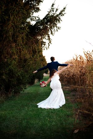 A couple in wedding attire dance joyfully on a grassy path between evergreen trees and autumn foliage at sunset.