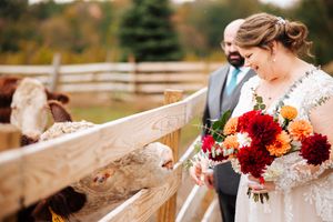 A couple in wedding attire share a joyful moment at a rustic farm fence while holding a vibrant red and orange bouquet at the Stables.
