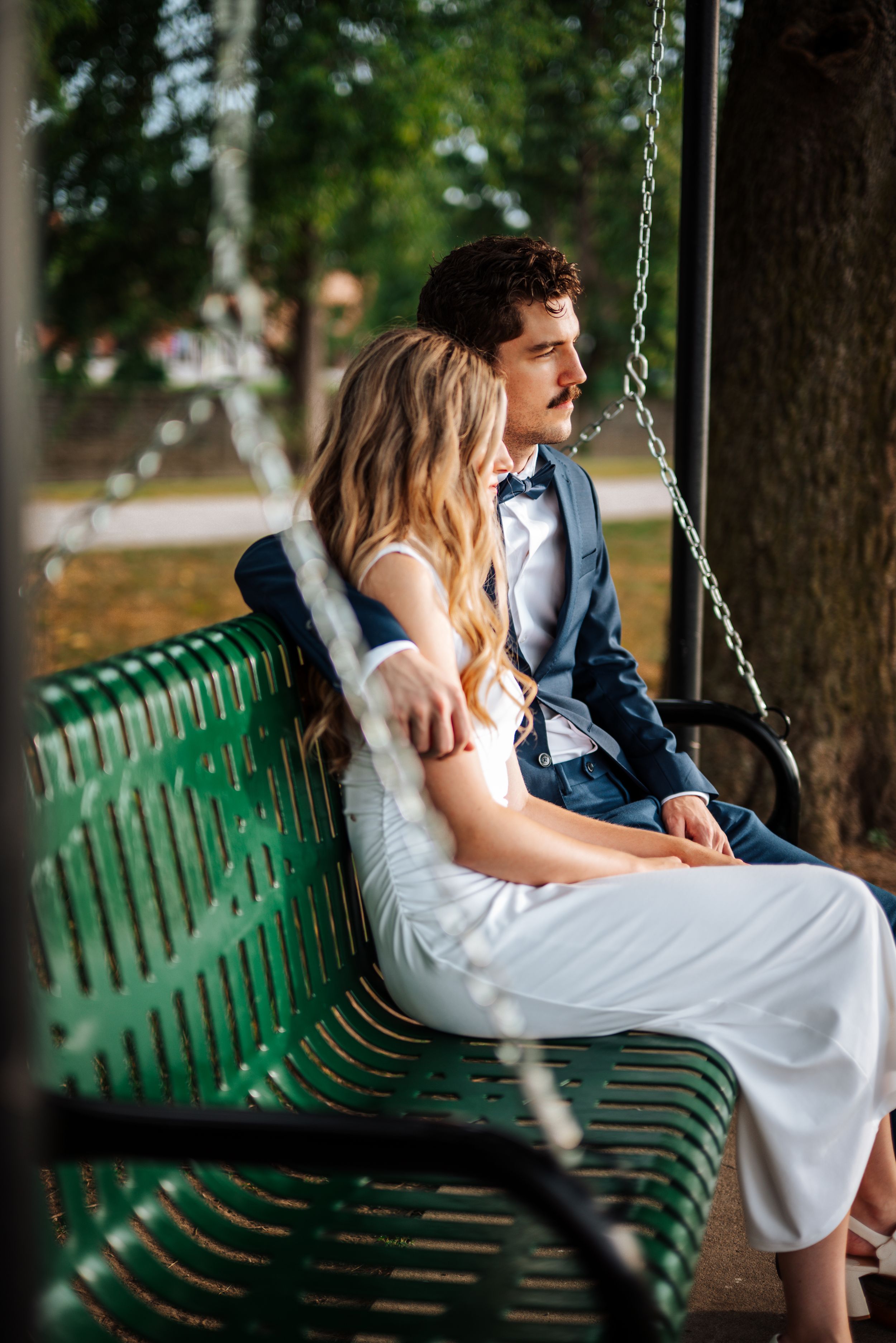 A couple in wedding attire sits closely on a green park bench swing, surrounded by lush trees.