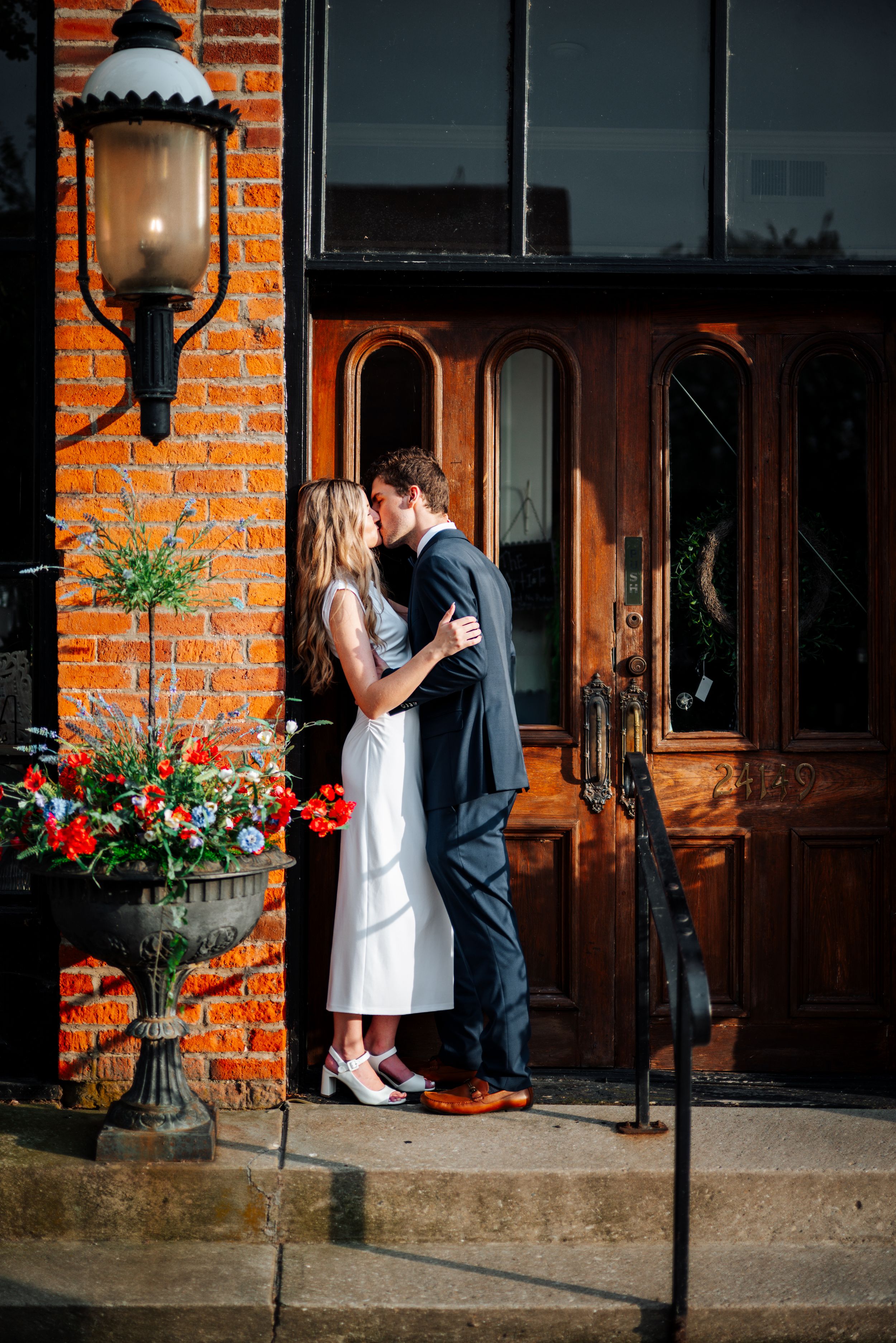 Couple sharing a romantic kiss on brick building steps beside wooden doors, flowers, and vintage lantern.