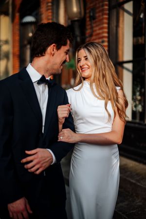 A smiling bride in a white gown and groom in a black tuxedo share a romantic moment outdoors.