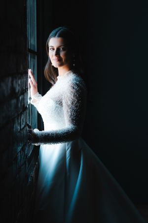 Elegant bride in beaded white gown standing by dark window, bathed in soft light.