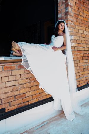 Elegant bride in white gown and veil posing against rustic brick wall, lifting dress to reveal heels.
