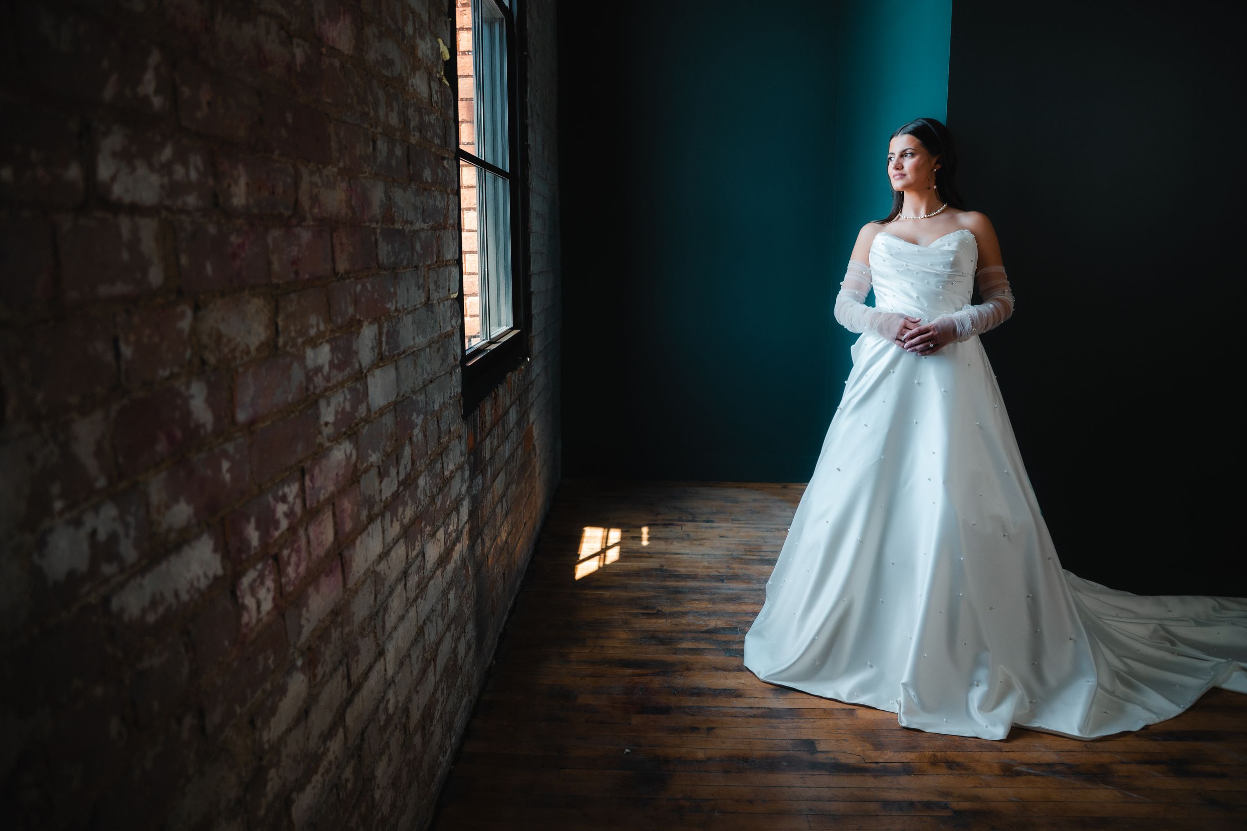 Bride in elegant white ball gown with long train stands by window in moody room with brick wall and teal accent.
