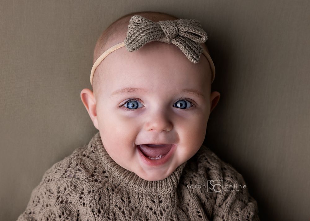 A happy baby wearing a gray knitted sweater and matching bow headband smiles brightly at the camera.