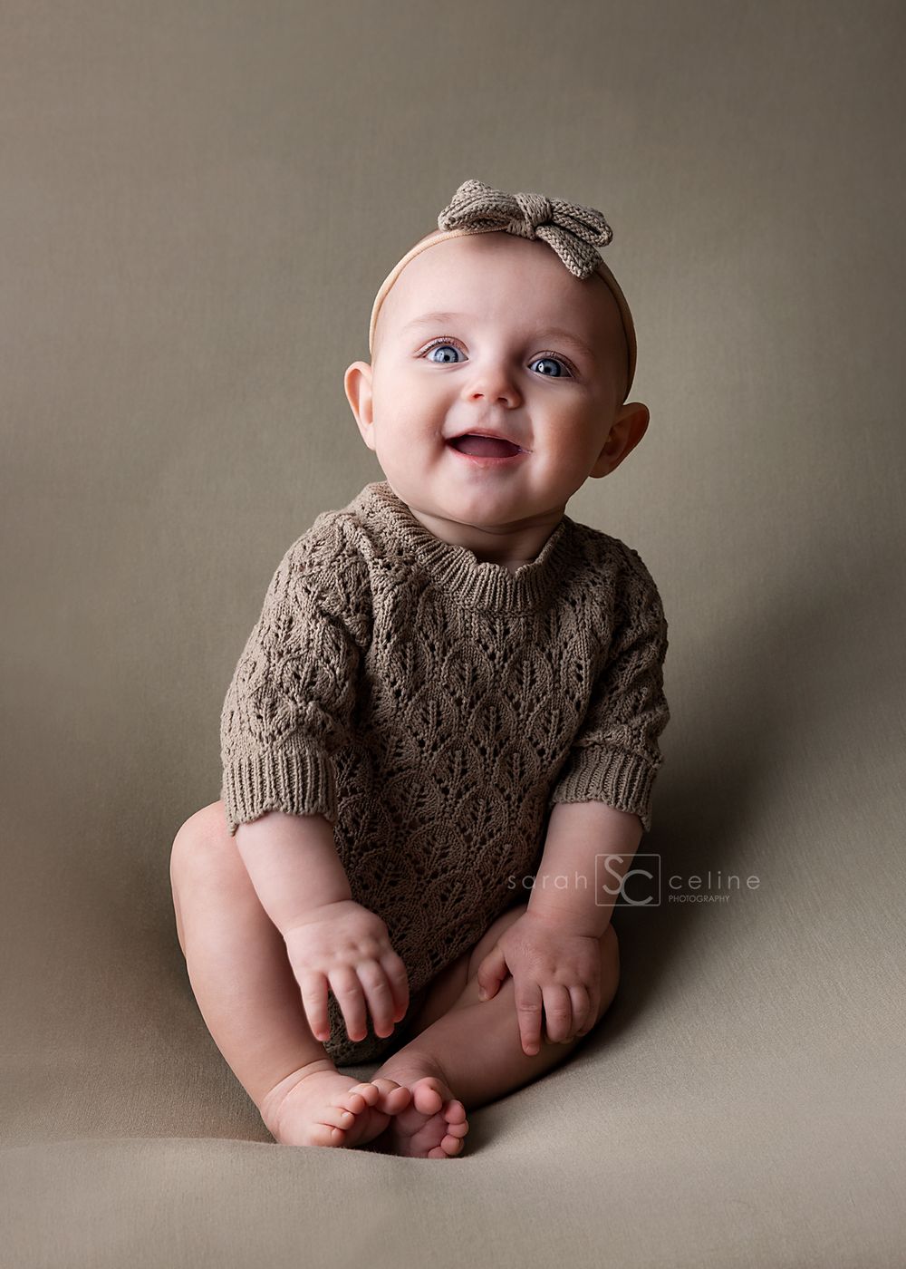 Baby wearing brown knit sweater and bow sits on neutral backdrop while smiling at camera during portrait session.