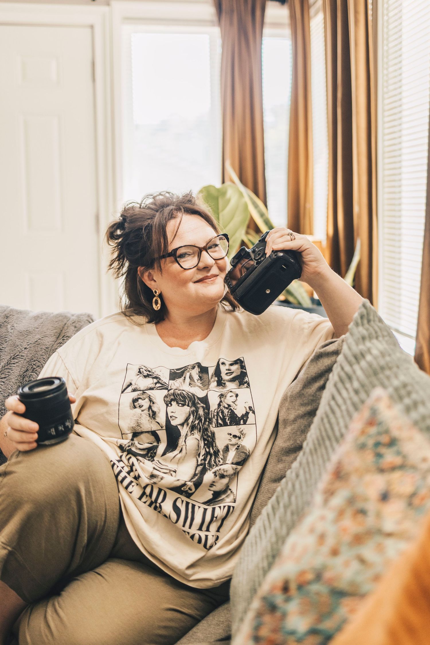 Woman in Taylor Swift shirt sitting on couch holding camera and lens.