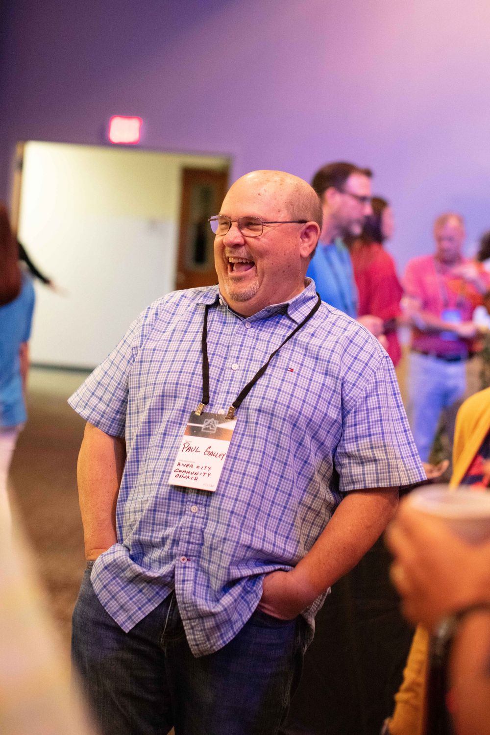 A person in a purple checkered shirt laughs and smiles during what appears to be a social gathering or event.