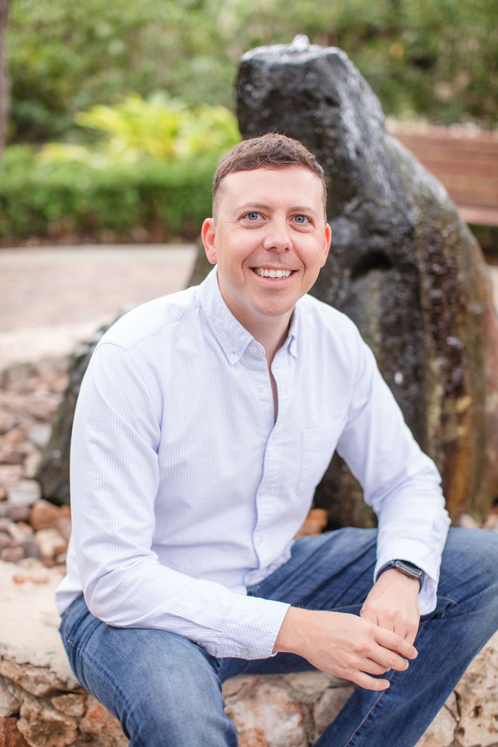 Professional portrait of someone in a white button-down shirt seated outdoors near a stone sculpture in a garden setting.