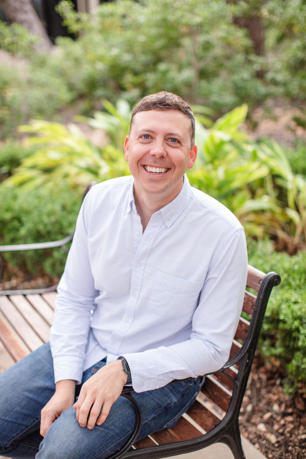 Casual outdoor portrait of someone in white shirt sitting on a wooden bench surrounded by lush greenery.
