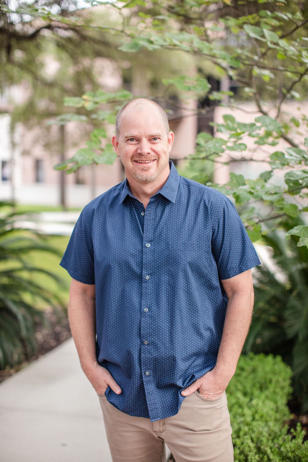 Outdoor portrait of someone wearing a blue short-sleeve button-down shirt and khakis standing on a garden path.