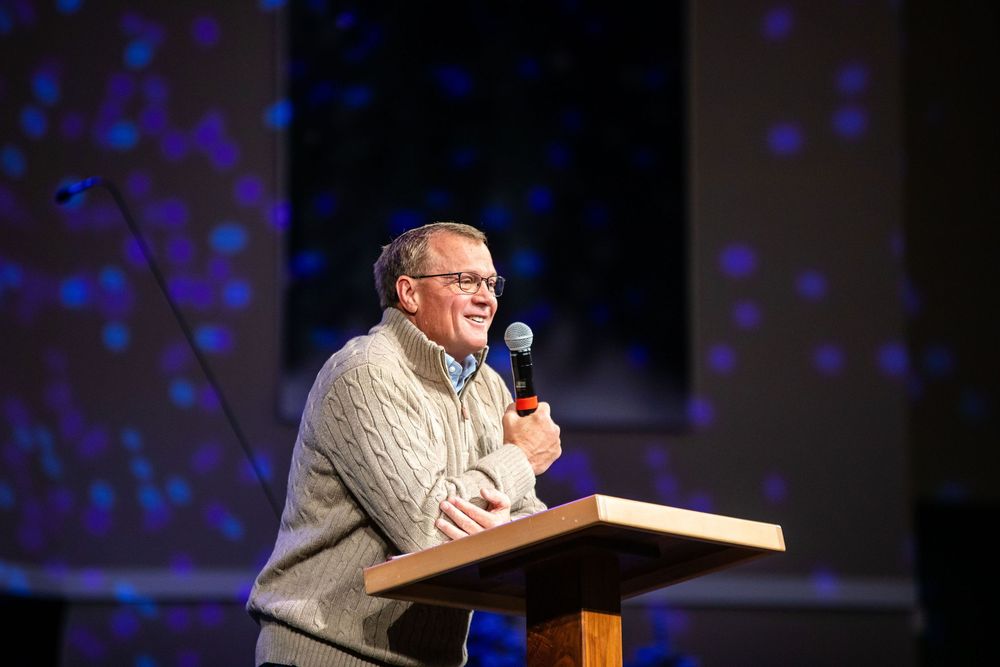 Speaker at a podium gestures passionately during presentation with blue lights in background.