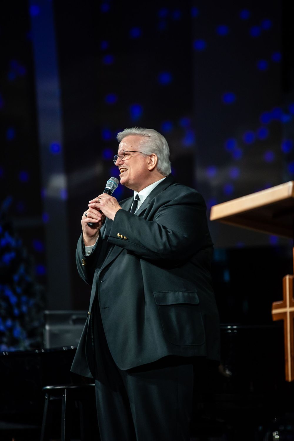 A performer in a black suit sings passionately on stage with blue lighting effects in the background.