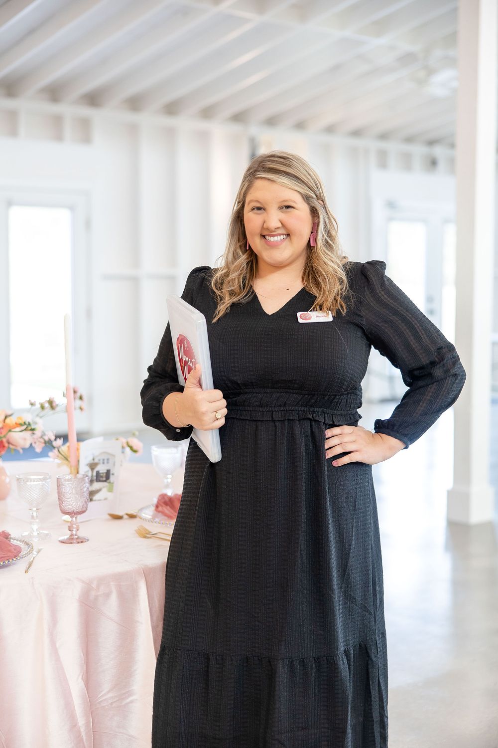A person wearing a long black dress poses and smiles in a bright white event space with pink table settings.