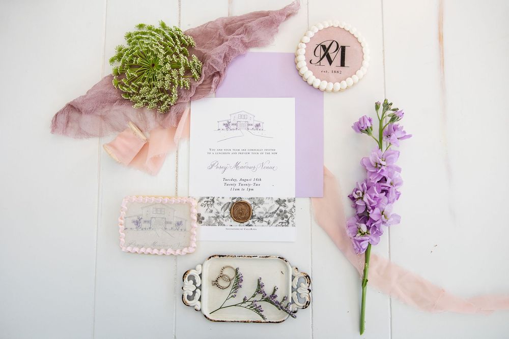 Wedding invitation flatlay with purple flowers, green succulent, pink ribbon, and decorative elements on white background.