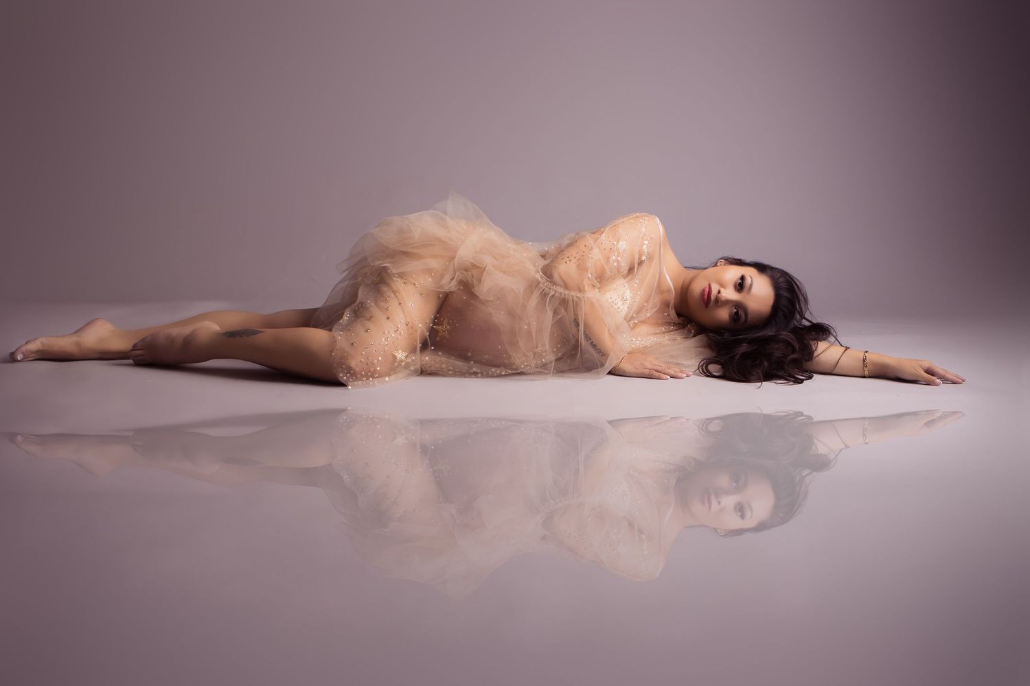 A graceful figure in a sheer beige dress lies reclined on a reflective studio floor with dramatic lighting.