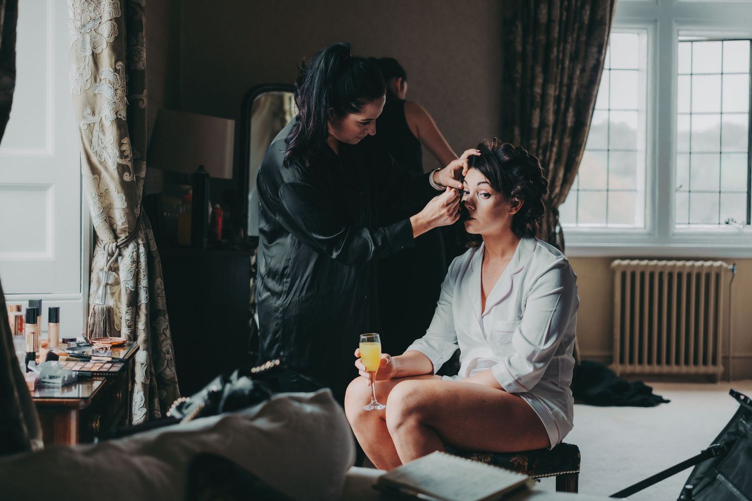 Makeup artist applies cosmetics to someone wearing a gray robe while sitting near a window in a vintage-style room.