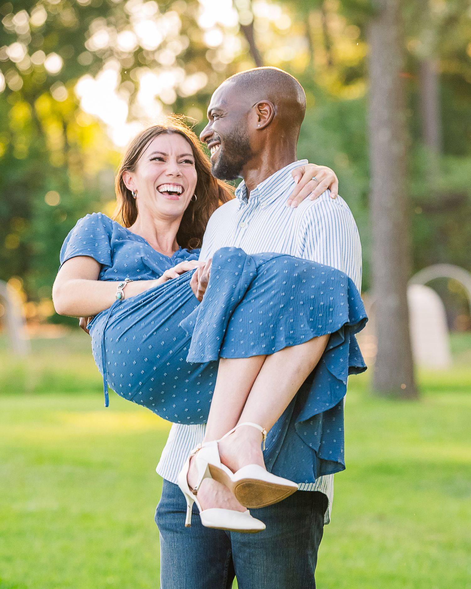 A joyful man lifts a laughing woman in a blue dress outdoors in a sunny park setting.