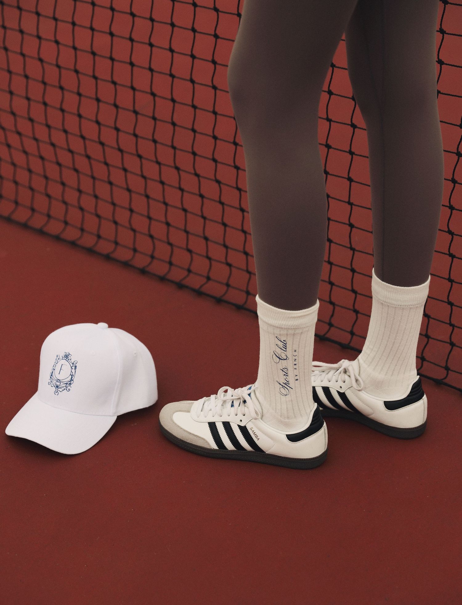 White baseball cap and classic Adidas sneakers against black tennis net and red court surface.