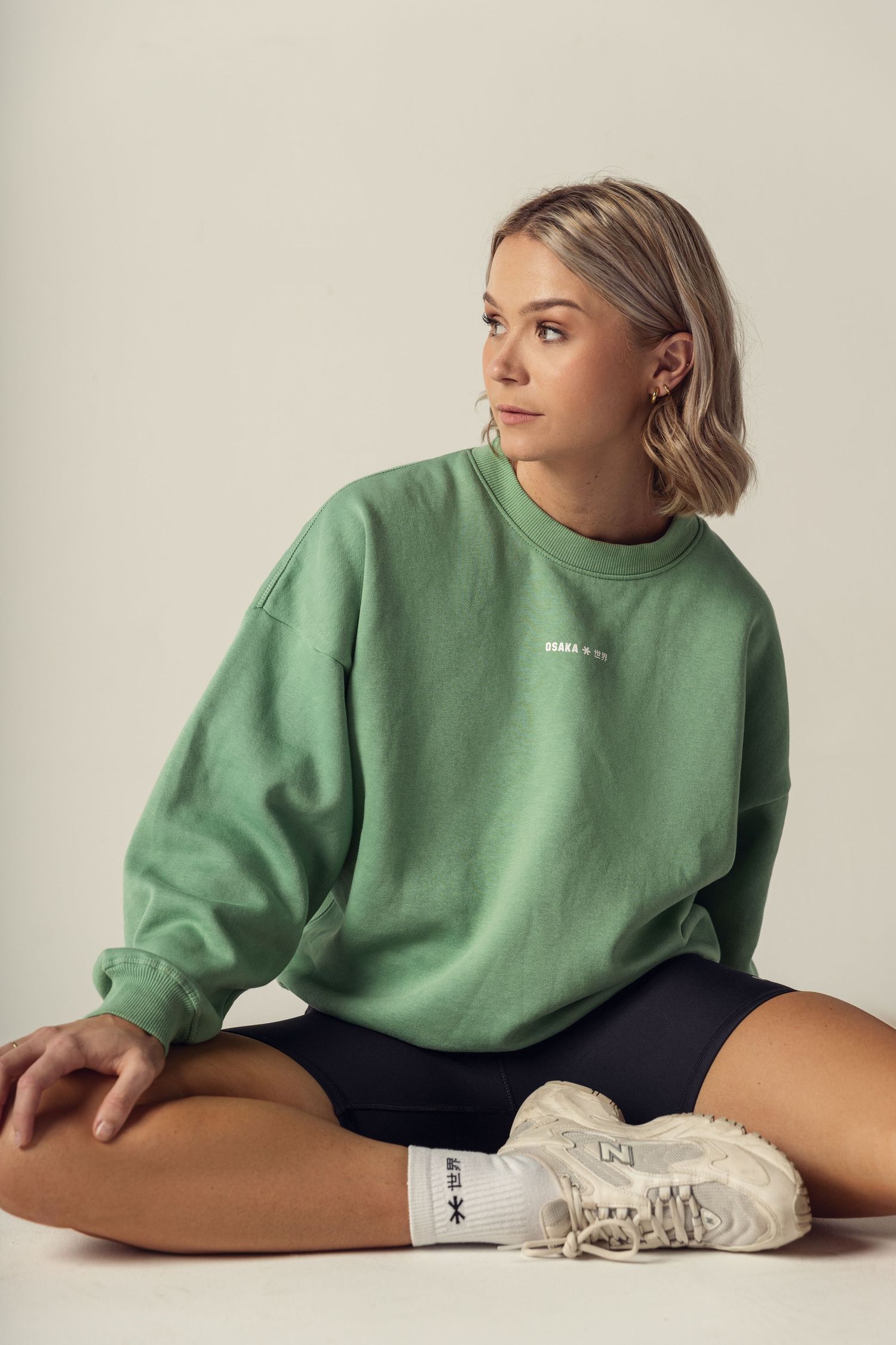 Athletic model in sage green sweatshirt and dark shorts sits cross-legged wearing white sneakers against neutral backdrop.